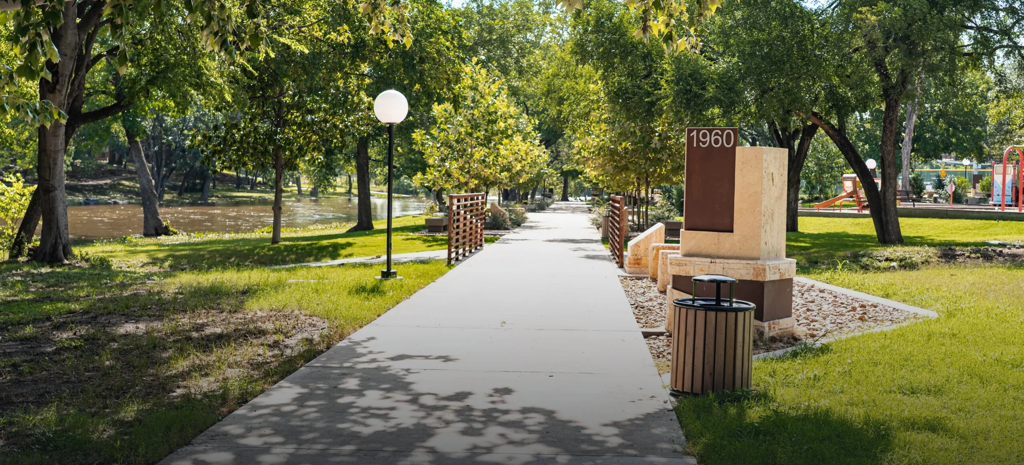 A shaded concrete trail running beside the creek in Memorial Park, Round Rock, with a playground beyond the trees