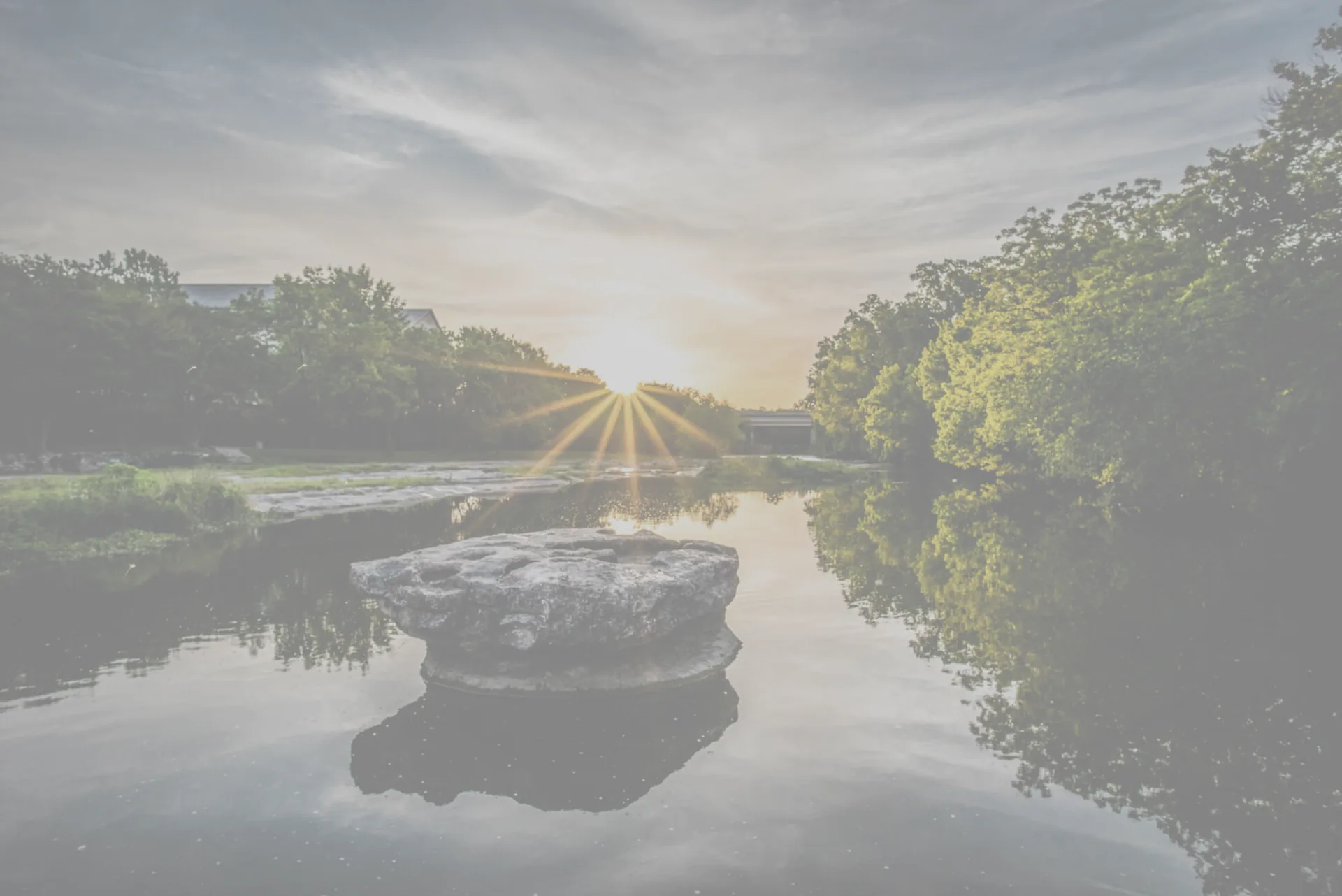 The round rock in Brushy Creek at sunrise, the sun breaking through the trees behind it and the whole scene mirrored in the water