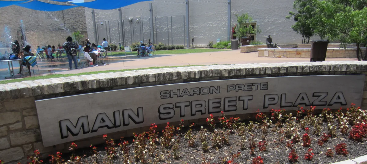 The Sharon Prete Main Street Plaza sign in downtown Round Rock, with the plaza tables and water feature behind it