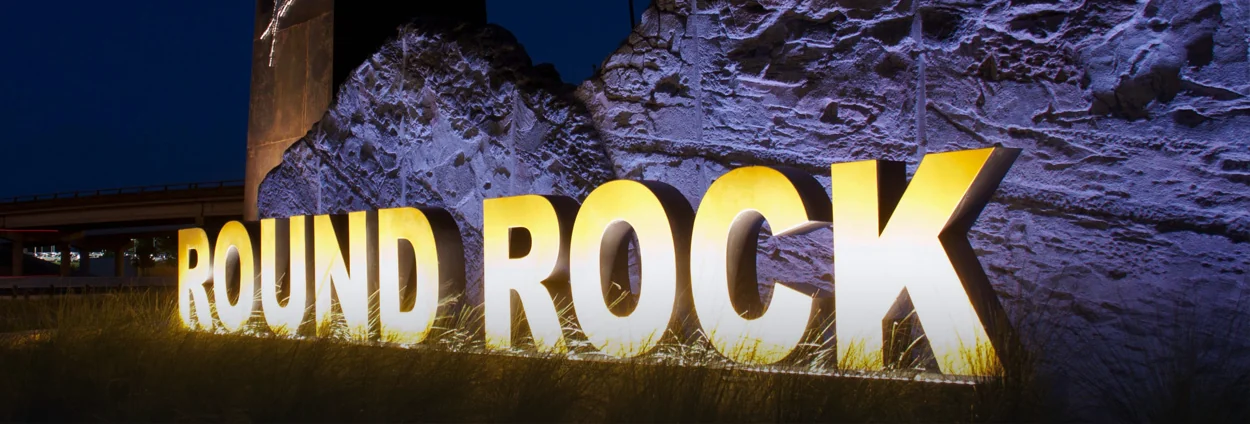 The lit ROUND ROCK sign at the city gateway at dusk, standing against a limestone cut