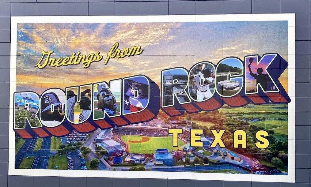 A Greetings from Round Rock, Texas postcard mural painted on a wall, with Dell Diamond and a Round Rock Express player inside the letters